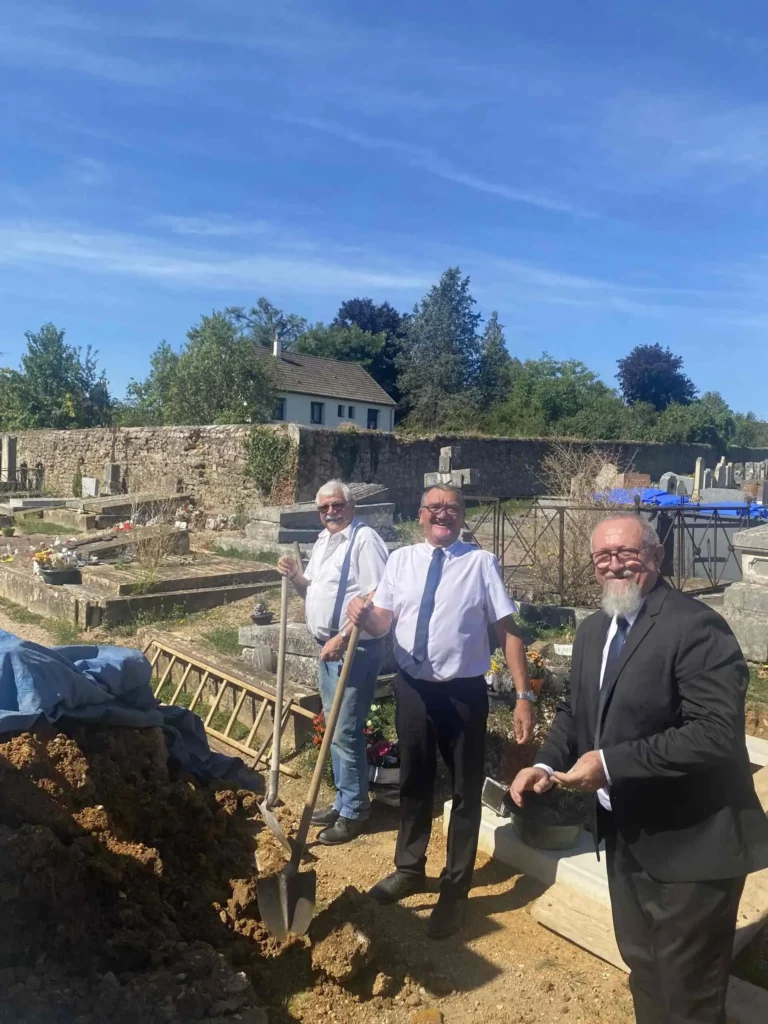 Trois employés funéraires souriants avec pelles devant une tombe ouverte dans un cimetière.