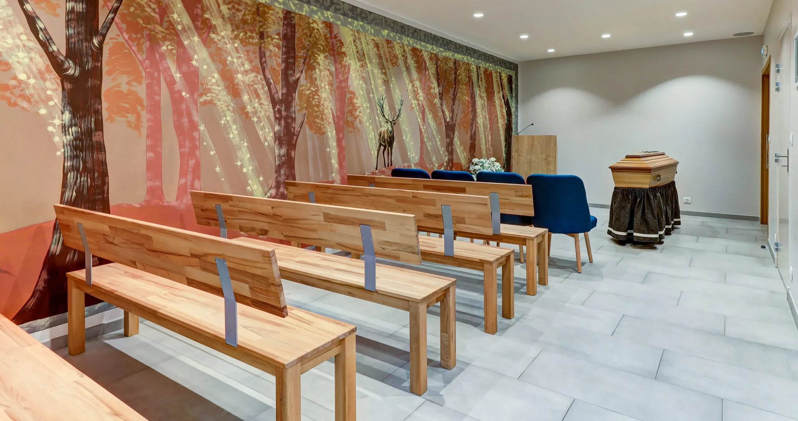 Salle de cérémonie funéraire avec bancs en bois et fresque murale de forêt.